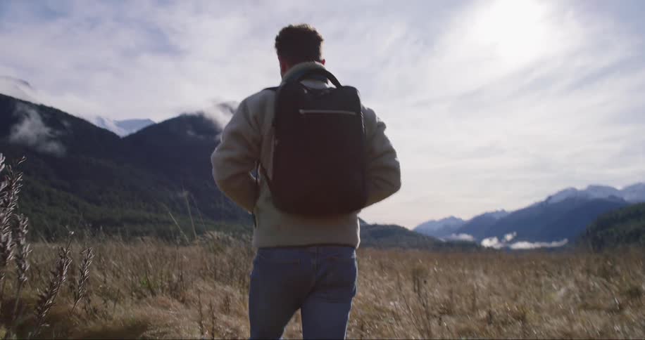 A man wearing a jacket and carrying a backpack is walking in a withered grassland with a mountain in sight