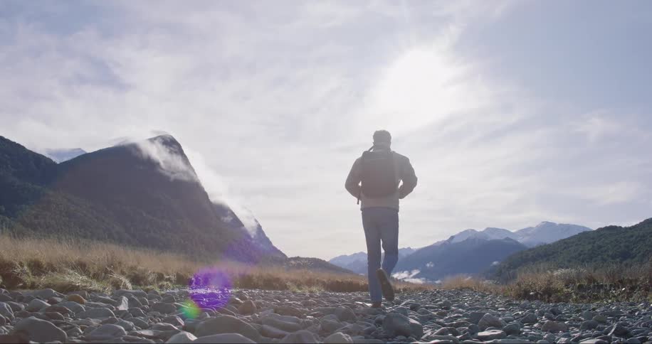 A man in a backpack and coat walks along a stone road in the middle of nature near the mountains