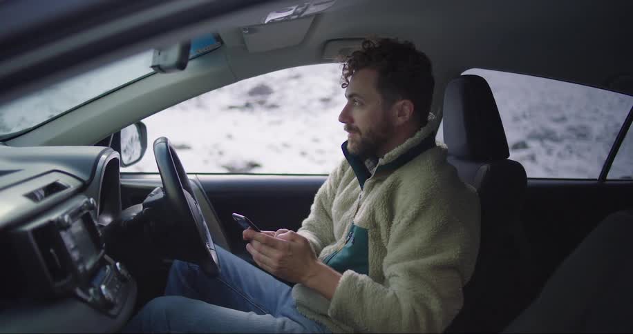 A man sitting in the driver seat of his parked car while texting on his phone and looking up periodically