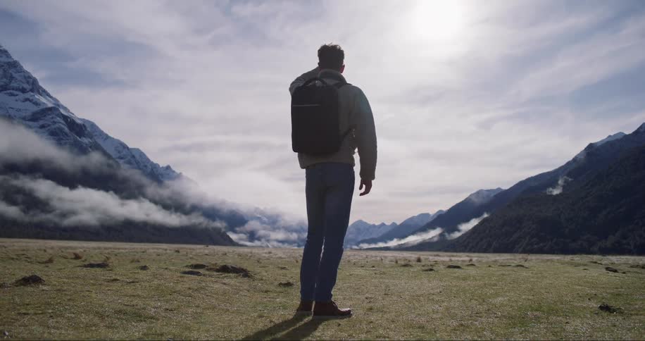 A man wearing a backpack stands and looks upon the mountainous landscape