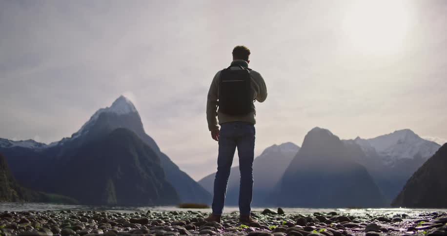A man in a backpack and jacket on the edge of a lake looking at the mountains and horizon
