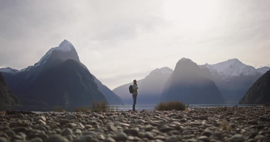 Man wearing backpack stands on rocky shore framed by mountains