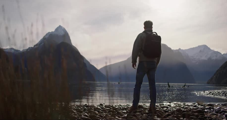 A hiker standing on a rocky lake shore looking around at the low mountain peaks around the lake on a hazy clouded fall day