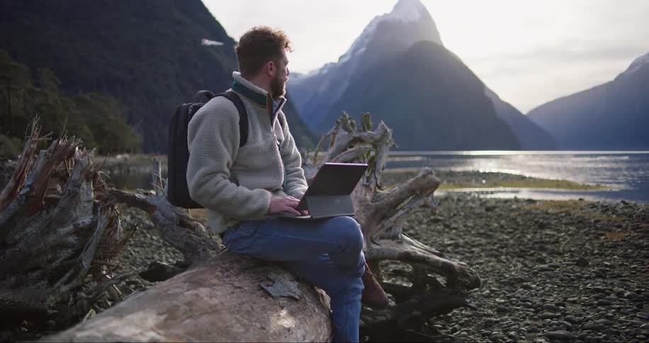 Man typing on tablet while sitting on fallen tree turns to watch plane fly between mountains and over water
