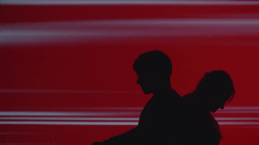 A male and a female silhouette pushing their backs together move into a standing position in front of an abstract red background with neon stripes