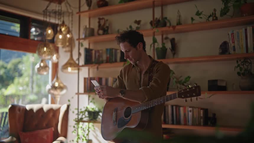 A man wearing a guitar indoors types on a smartphone and contemplates