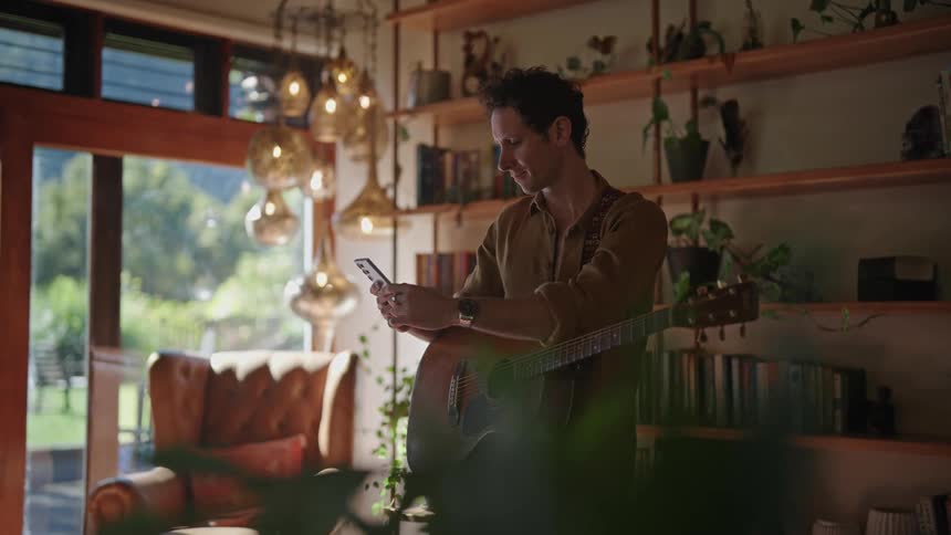 Young man holding an acoustic guitar browses his phone in the living room
