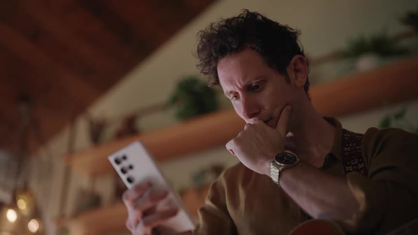 A man with dark hair touches his face and looks at his phone while standing near a bookshelf