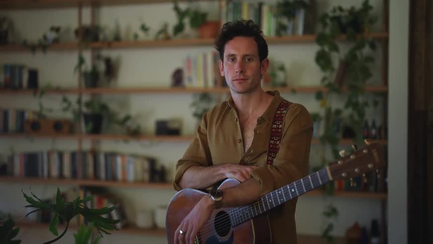 Man with dark hair holding a guitar standing in front of a bookshelf