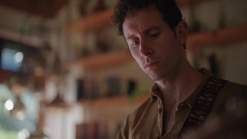 Man in brown shirt wristwatch and ring plays a guitar looking down