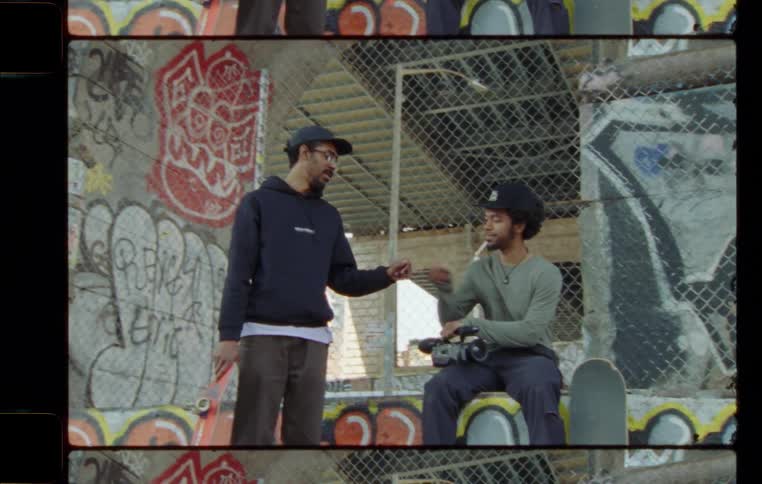 A man of the skateboard talks to and leaves a man sitting on a cement wall with a camera under a bridge filled with graffiti