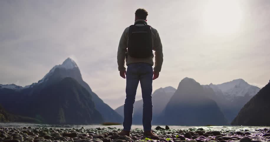Man wearing backpack stands on rocky coast with mountains ahead