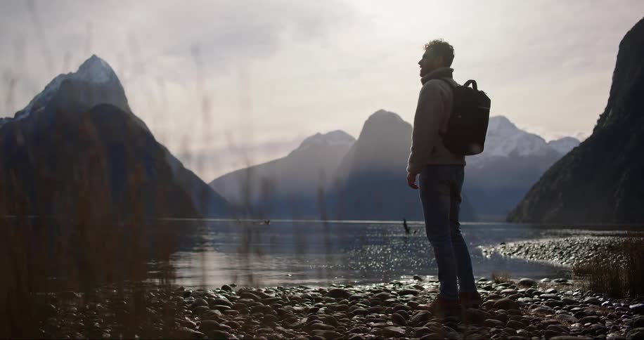 A man in a coat and backpack is standing on the side of a lake with mountains in the background