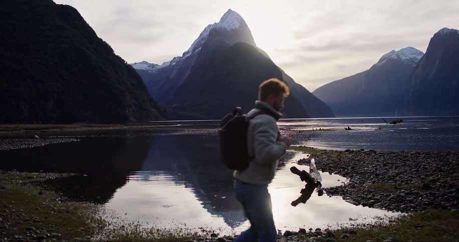 A hiker walking by water in front of a mountain landscape on a cool day