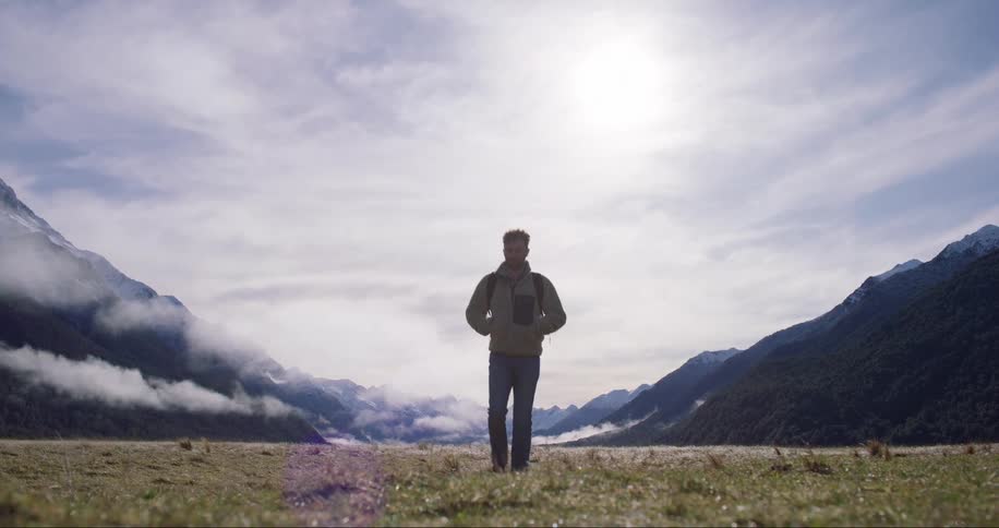 A man with hands in his pockets walking straight ahead and in the background are big mountains