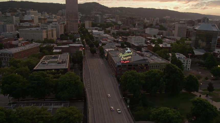 Aerial footage captures the beautiful sunset over downtown Portland Oregon as the drone follows the Willamette River towards the iconic White Stag Sign showcasing the historical high-rise buildings and famous landmarks of the city