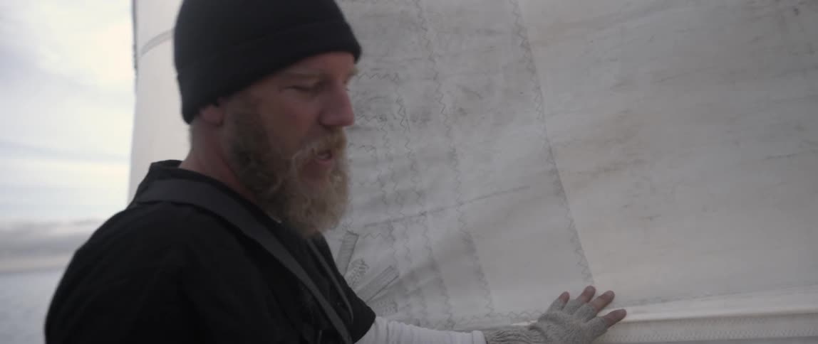 A man standing near a sail on a boat instructs is pointing at someone