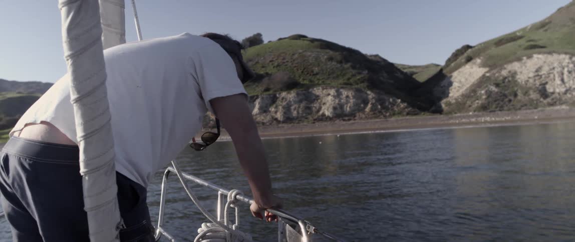 A man is leaning over the side of a boat by the shore
