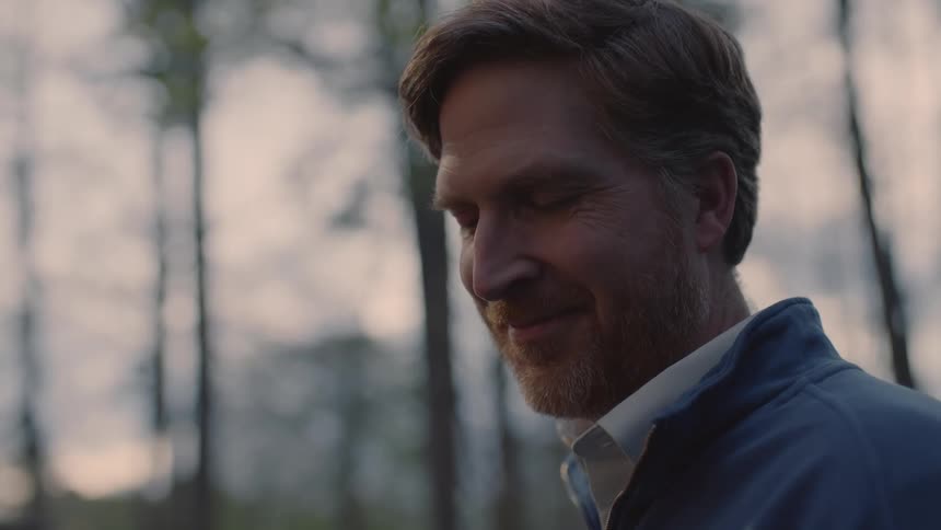 A man stands smiling in a forest with tall trees