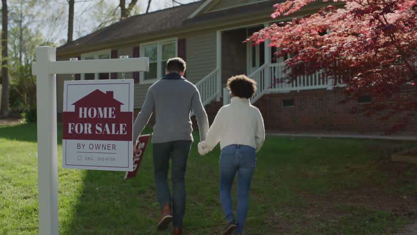 White man and black woman walk towards a sign and remove the part written sold before walking towards the house during day time