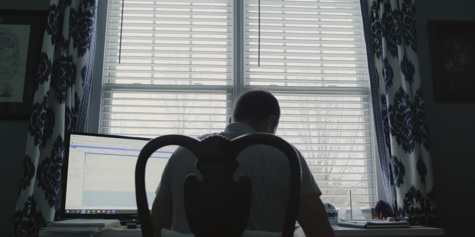 A man sitting in a chair reading a paper next to a computer screen