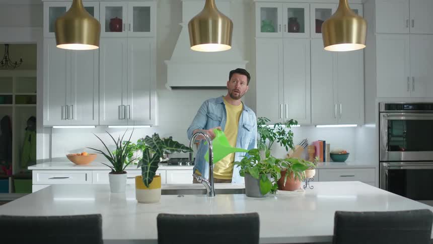 A man in a white kitchen watering plants that are on the table