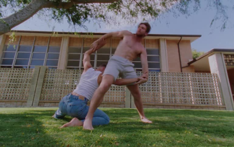 Two men are practicing acrobatic dance moves in the front yard of a building one is wearing jeans and a singlet while the other is wearing just shorts a tree and grass are also visible in the background