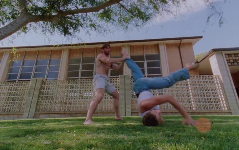 Two male dancers performing on the grass outdoors under a tree next to a building during day time