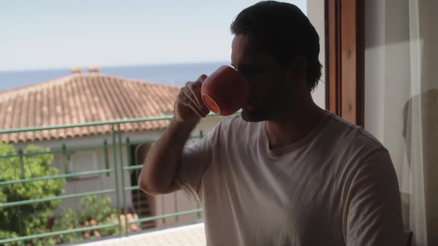 A man sitting at a table near a villa balcony takes a sip from a mug while typing on a laptop