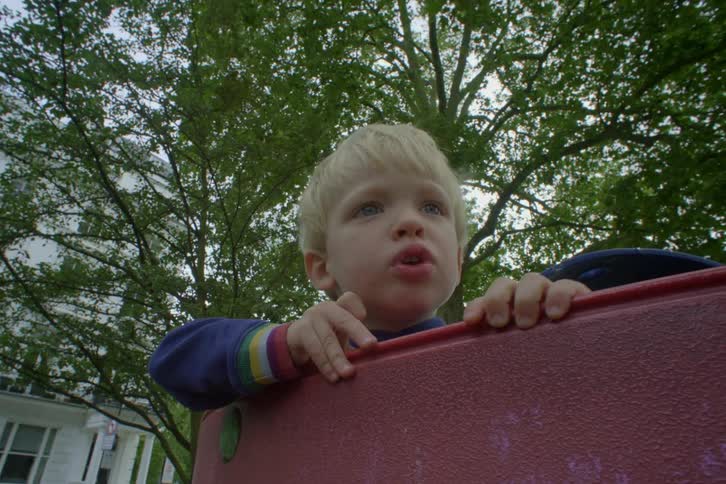 A kid in the playground surrounded by trees and a white house on the left