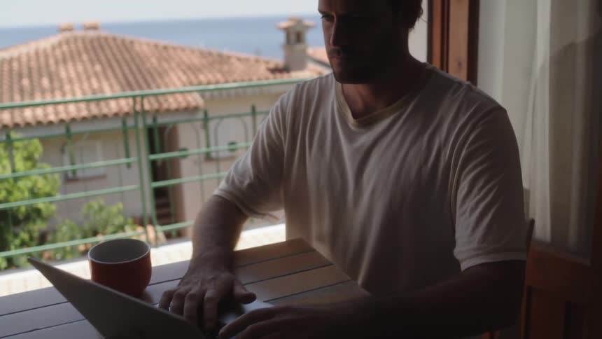 A man sitting at a table during a bright sunny day while he types on his laptop in front of him