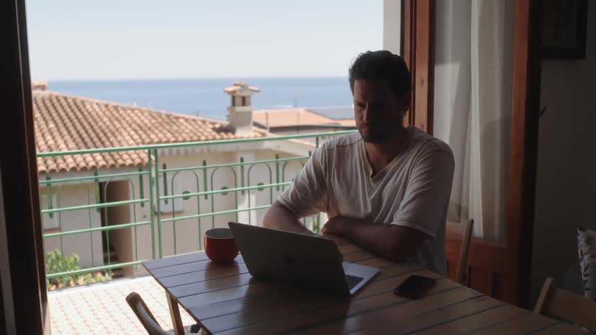 A man sitting at the table observing the ocean view and working on his laptop
