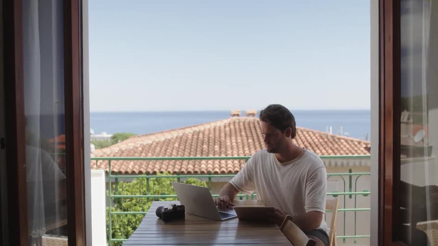 A man sitting at a table on a balcony during the day while using his laptop and writing