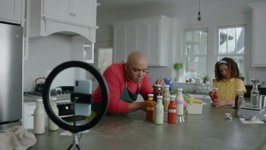 A teenage daughter putting labels on a jar while her father places jars in front of a camera and light in their kitchen during the day