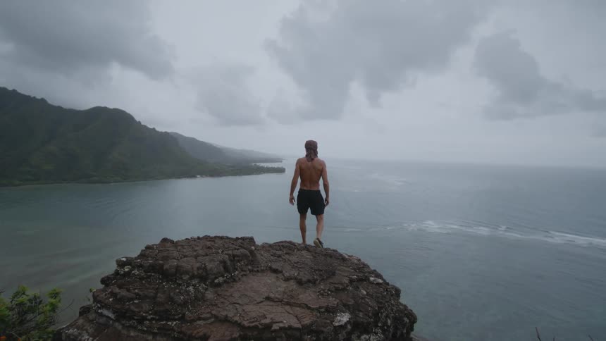 Young shirtless man stands on the edge of a cliff in front of the ocean