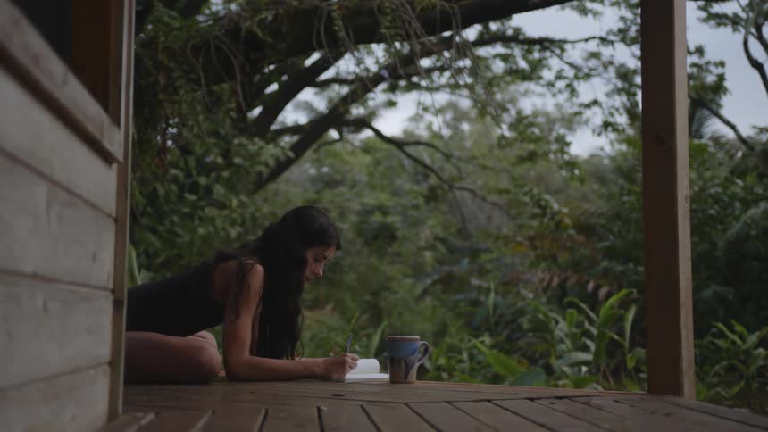 A woman lying on the floor of her balcony in her backyard while writing on a book with a coffee mug on her side with trees surrounding the building