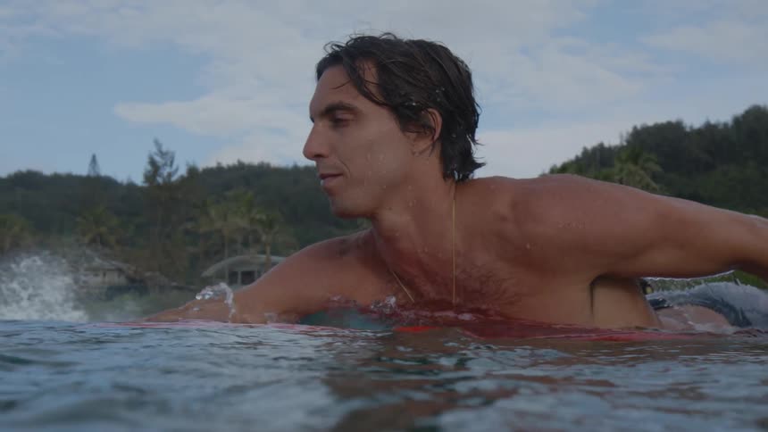 Young shirtless man swims over a surfboard near the coast