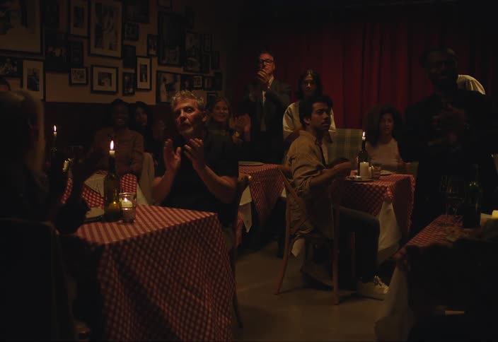 A small audience sits at tables in a dimly lit room and clap