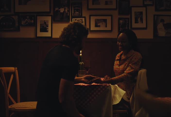 A white man and a black woman are in a restaurant theres a glass of wine and a candle on the table suddenly the man goes down on one knee to propose to the lady and they hug afterward