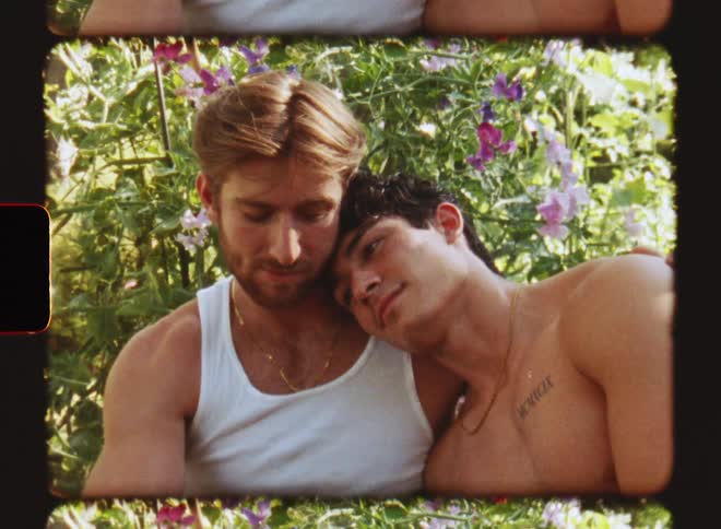 Two men cuddling outdoors during the day in front of a flowering plant one without a shirt on