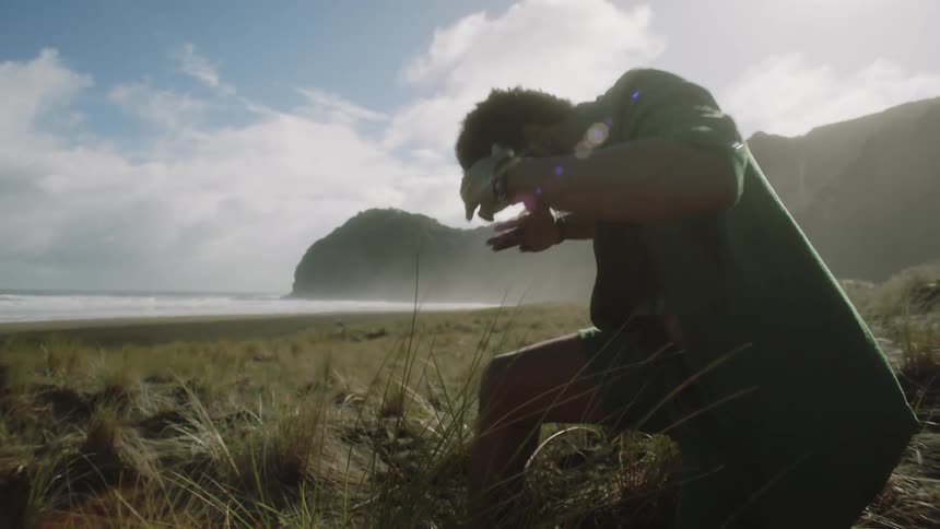 A black man is kneeling on one foot on the coastline near the beach surrounded by grasses he is using his hands to catch the wind in a form of religious activity