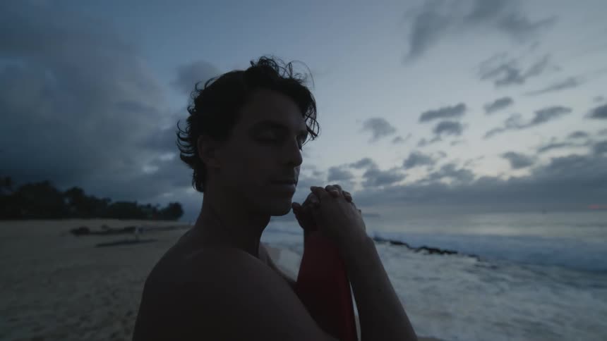 A young man on the beach sand facing the sea holding his surfboard