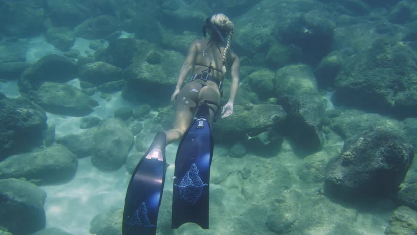 A woman snorkeling and swimming underwater near rocks on the ocean floor as she spins around