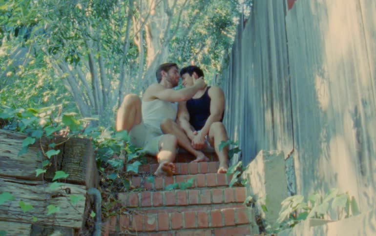 Two men kiss while sitting on brick steps under a tree outdoors