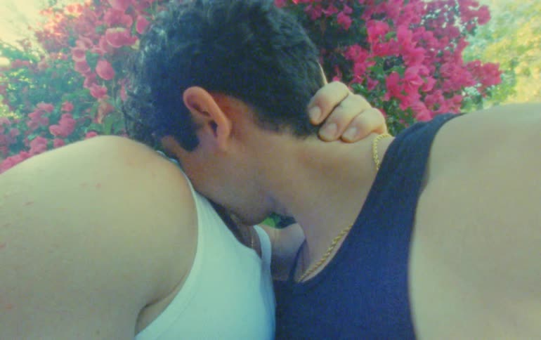 Two men hugging cuddling and kissing each other while standing in front of a pink flowering bush during a sunny day
