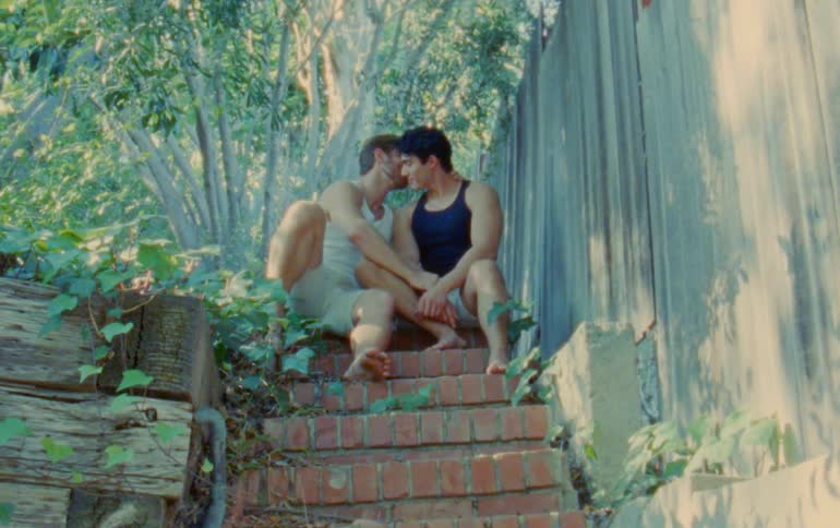 Two men sitting outdoors on brick steps are kissing underneath some trees