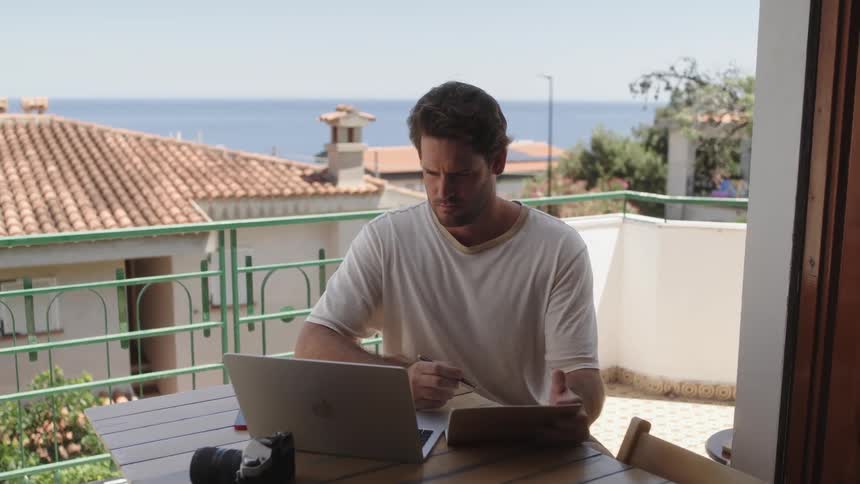 A man working with a notebook sits at a table on the porch