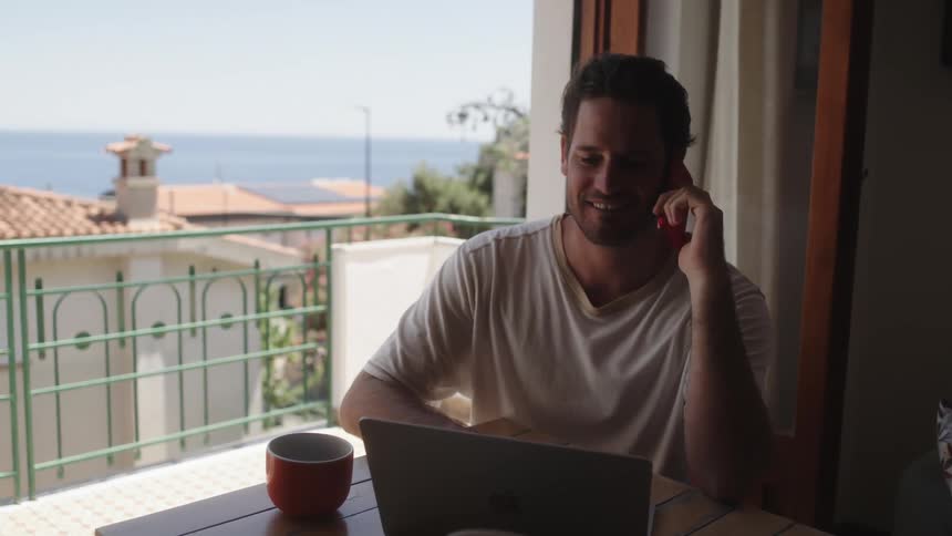 A man with a laptop sitting by a window with a balcony talking on the phone and smiling