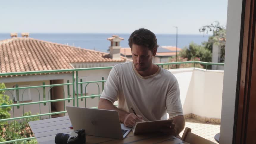 A man sitting on the balcony writing in a notebook and glancing at the laptop beside him