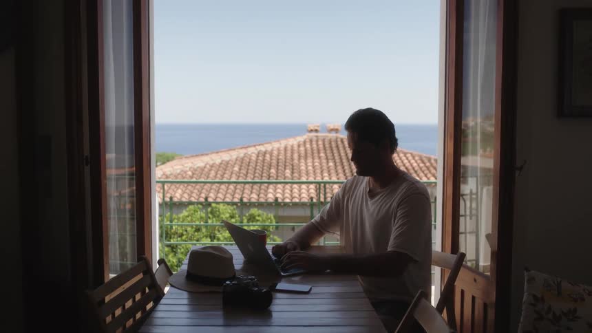 Man sits at table working on laptop in front of open doors